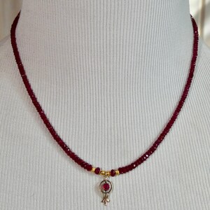 May include: A delicate necklace featuring a strand of small, deep red beads. A small pendant with a red gemstone and gold accents hangs from the center. The necklace is displayed on a white mannequin.