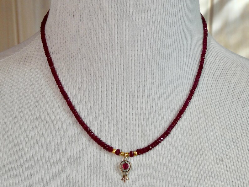 May include: A delicate necklace featuring a strand of small, deep red beads. A small pendant with a red gemstone and gold accents hangs from the center. The necklace is displayed on a white mannequin.