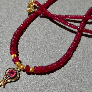 May include: A ruby necklace with a gold pendant. The necklace features small, faceted ruby beads strung together. The pendant is gold and has a circular ruby stone in the center. The necklace is displayed on a gray surface.