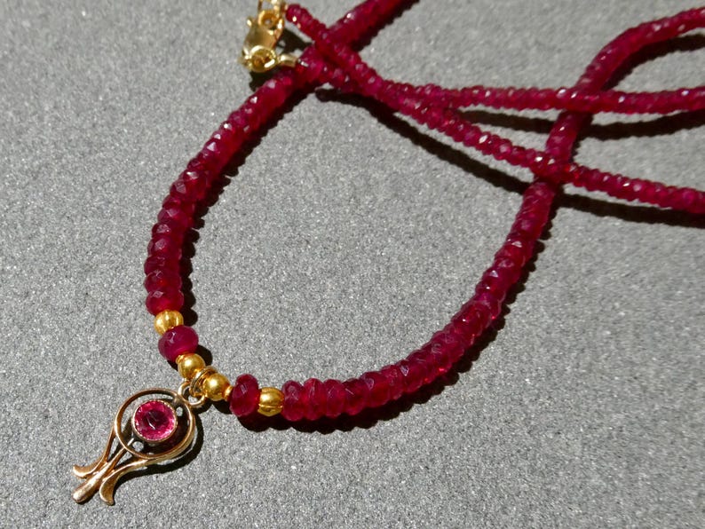 May include: A ruby necklace with a gold pendant. The necklace features small, faceted ruby beads strung together. The pendant is gold and has a circular ruby stone in the center. The necklace is displayed on a gray surface.
