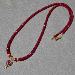 May include: A ruby necklace with a gold pendant. The necklace is made of small, faceted red beads. The pendant is gold with a small, round ruby in the center. The necklace has a gold clasp.