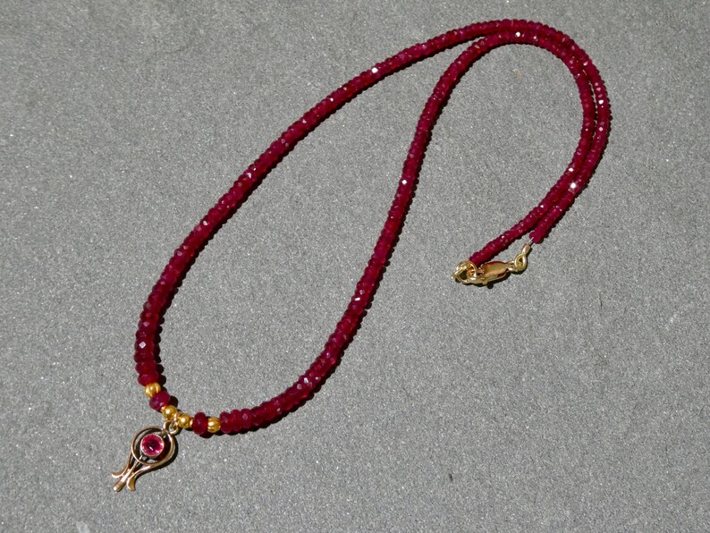 May include: A ruby necklace with a gold pendant. The necklace is made of small, faceted red beads. The pendant is gold with a small, round ruby in the center. The necklace has a gold clasp.