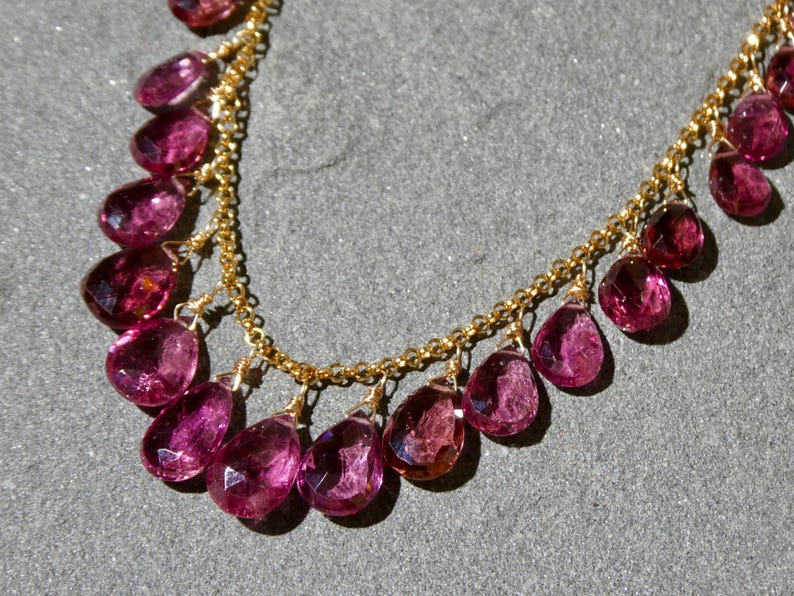 May include: A necklace featuring a delicate gold chain adorned with numerous faceted, teardrop-shaped magenta gemstones. Each stone is individually wire-wrapped and suspended from the chain, creating a cascading fringe effect along the neckline.