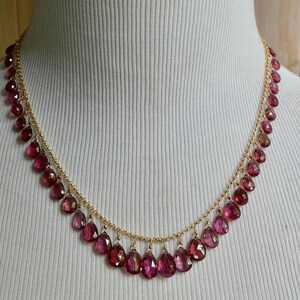 May include: A gold chain necklace featuring a series of dangling, faceted, teardrop-shaped dark pink gemstones. The stones are evenly spaced along the length of the delicate gold chain, creating a fringe-like effect around the neckline.