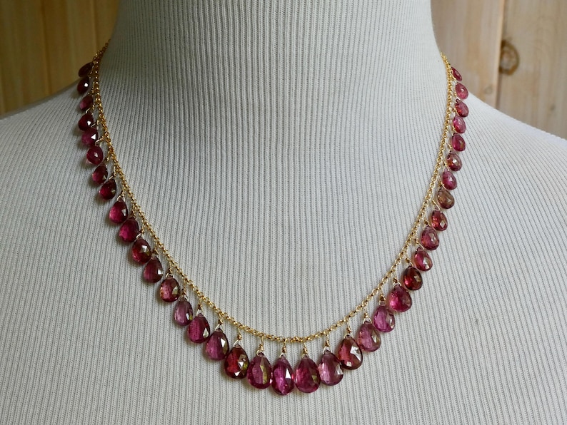 May include: A gold chain necklace featuring a series of dangling, faceted, teardrop-shaped dark pink gemstones. The stones are evenly spaced along the length of the delicate gold chain, creating a fringe-like effect around the neckline.