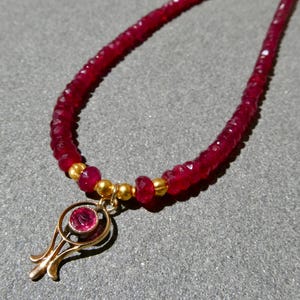 May include: A ruby necklace featuring a strand of small, faceted red beads. The necklace has a gold-colored pendant with a central ruby stone. The pendant has a stylized, openwork design. The necklace is displayed on a gray surface.