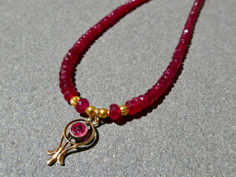 May include: A ruby necklace featuring a strand of small, faceted red beads. The necklace has a gold-colored pendant with a central ruby stone. The pendant has a stylized, openwork design. The necklace is displayed on a gray surface.