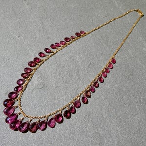 May include: A gold chain necklace featuring a series of faceted, teardrop-shaped magenta gemstones suspended along the length of the chain. The stones are arranged in a graduated pattern, with the largest gems positioned at the center of the necklace.