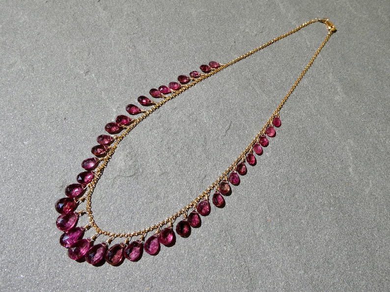 May include: A gold chain necklace featuring a series of faceted, teardrop-shaped magenta gemstones suspended along the length of the chain. The stones are arranged in a graduated pattern, with the largest gems positioned at the center of the necklace.