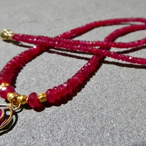 May include: A close-up of a ruby necklace with a gold pendant. The necklace features small, faceted ruby beads strung together, creating a vibrant red color. The pendant is gold with a ruby center. The necklace is displayed on a gray surface.