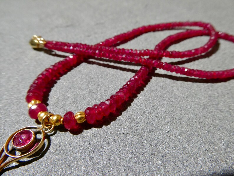 May include: A close-up of a ruby necklace with a gold pendant. The necklace features small, faceted ruby beads strung together, creating a vibrant red color. The pendant is gold with a ruby center. The necklace is displayed on a gray surface.