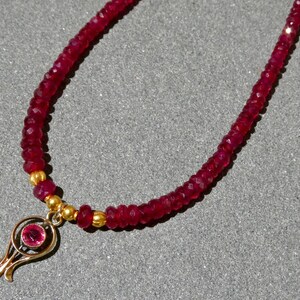 May include: A ruby necklace with a gold pendant. The necklace features small, faceted red ruby beads strung together. The gold pendant has a floral design with a central ruby. The necklace is displayed on a gray surface.