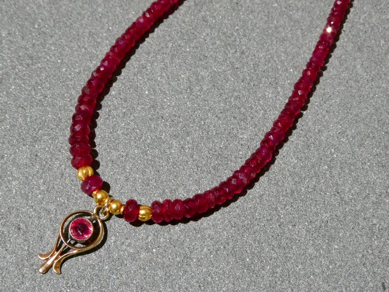 May include: A ruby necklace with a gold pendant. The necklace features small, faceted red ruby beads strung together. The gold pendant has a floral design with a central ruby. The necklace is displayed on a gray surface.
