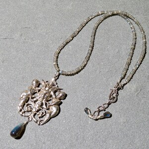 May include: A silver necklace with a pendant featuring a detailed design of multiple figures intertwined. The pendant is attached to a silver chain with a silver clasp. The necklace is made of light grey beads.