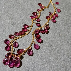 May include: A delicate gold chain necklace featuring numerous faceted, teardrop-shaped magenta gemstones suspended along the length of the chain. The necklace displays a cascading arrangement of these small, translucent pinkish-red stones.