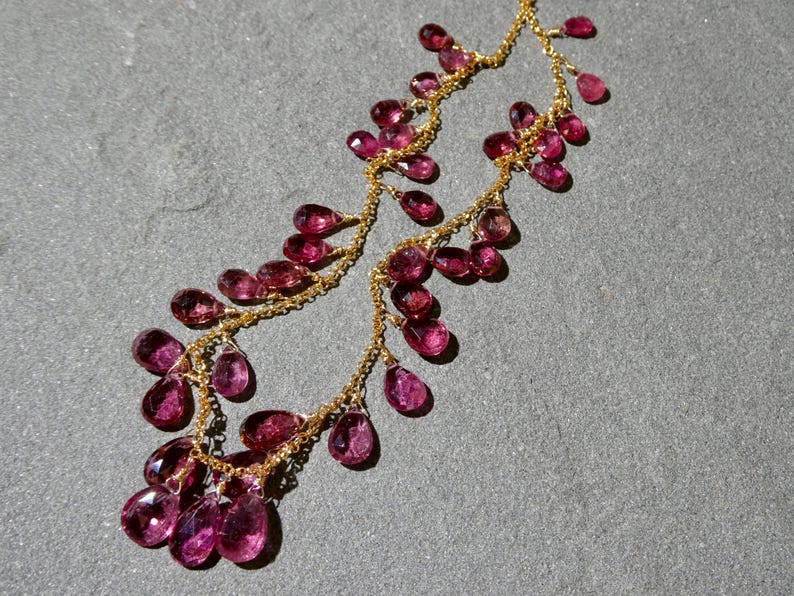 May include: A delicate gold chain necklace featuring numerous faceted, teardrop-shaped magenta gemstones suspended along the length of the chain. The necklace displays a cascading arrangement of these small, translucent pinkish-red stones.