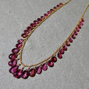 May include: A gold chain necklace featuring a series of faceted, teardrop-shaped magenta gemstones suspended along the length of the chain. The stones are arranged in a graduated pattern, with larger gems at the center and smaller ones toward the ends.