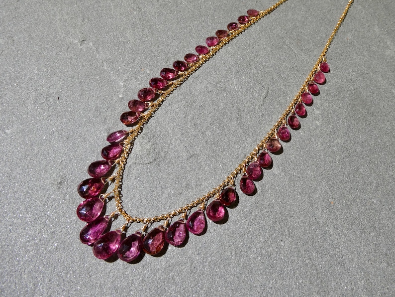 May include: A gold chain necklace featuring a series of faceted, teardrop-shaped magenta gemstones suspended along the length of the chain. The stones are arranged in a graduated pattern, with larger gems at the center and smaller ones toward the ends.