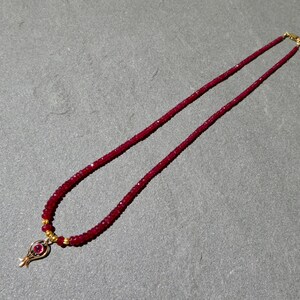 May include: A ruby red beaded necklace with a gold pendant. The necklace features small, round beads and a gold clasp. The pendant has a gold frame with a ruby red stone in the center. The necklace is displayed on a gray surface.
