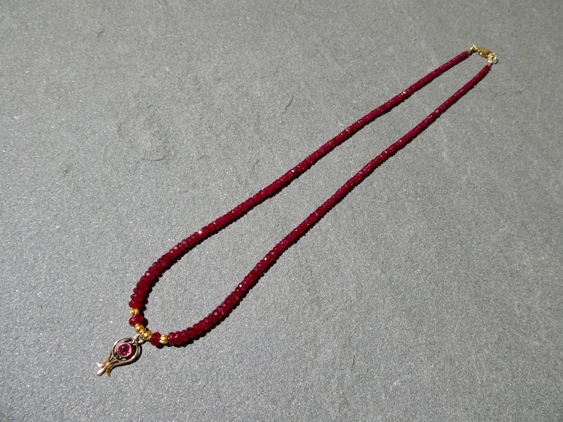 May include: A ruby red beaded necklace with a gold pendant. The necklace features small, round beads and a gold clasp. The pendant has a gold frame with a ruby red stone in the center. The necklace is displayed on a gray surface.