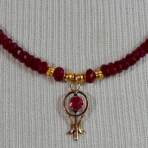 May include: A burgundy beaded necklace with a gold pendant. The pendant features a red gemstone set in a gold filigree design. The necklace is composed of small, faceted beads.