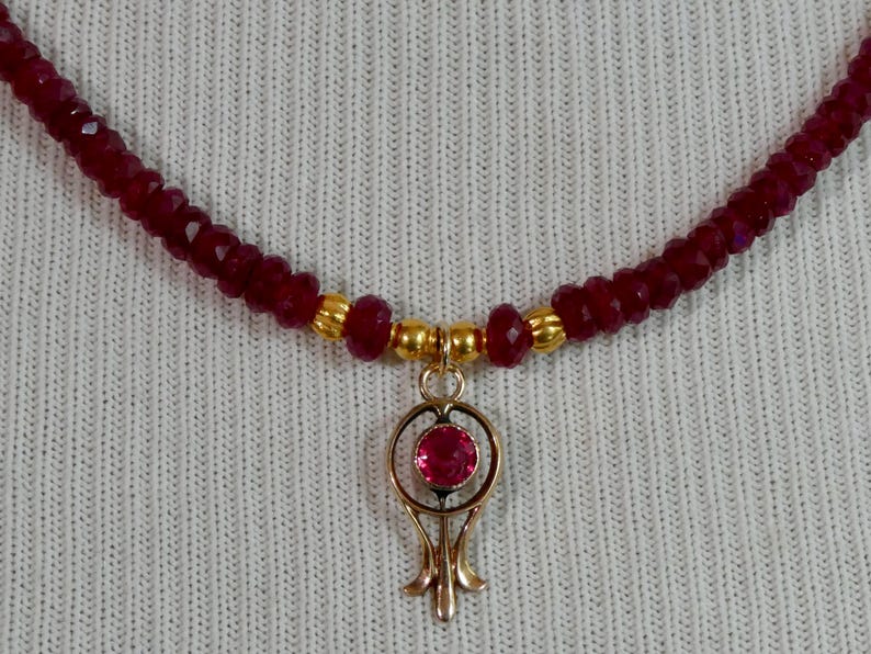 May include: A burgundy beaded necklace with a gold pendant. The pendant features a red gemstone set in a gold filigree design. The necklace is composed of small, faceted beads.