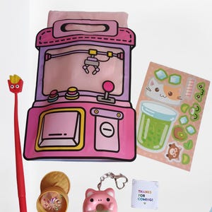 May include: A pink claw machine pouch, a red pen with a french fry topper, a sheet of stickers featuring a cat and drink, a small gold container with a clear lid, a pink donut keychain, and a small white card with the text THANKS FOR COMING!