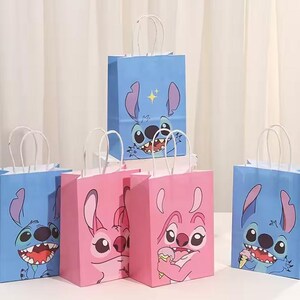 May include: A collection of blue and pink paper gift bags featuring cartoon character designs. The bags have white paper handles and are arranged on a white surface. The bags have a cartoon character's face printed on them.