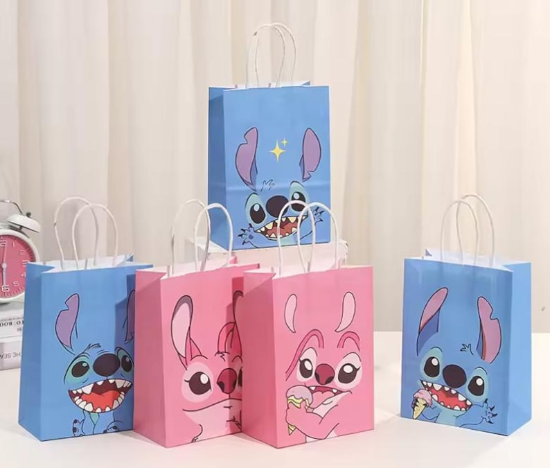 May include: A collection of blue and pink paper gift bags featuring cartoon character designs. The bags have white paper handles and are arranged on a white surface. The bags have a cartoon character's face printed on them.
