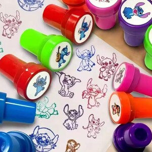 May include: A collection of colorful, cartoon character stamps with red, green, pink, orange, and purple handles. The stamps feature various images of a blue alien character. The stamped images are in blue, red, and black ink.
