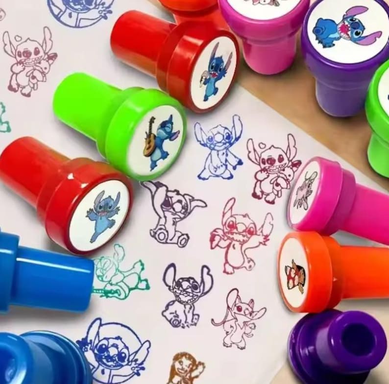 May include: A collection of colorful, cartoon character stamps with red, green, pink, orange, and purple handles. The stamps feature various images of a blue alien character. The stamped images are in blue, red, and black ink.