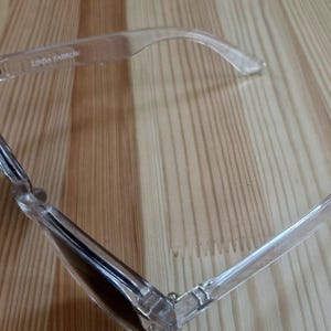 Peut inclure: Lunettes de soleil &agrave; monture transparente avec verres fonc&eacute;s, pos&eacute;es sur une surface en bois. Le nom de la marque "LINDA FARROW" est visible sur la monture. Les lunettes ont un design moderne et minimaliste.