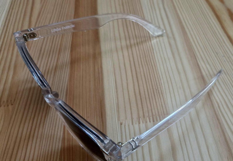 Peut inclure: Lunettes de soleil &agrave; monture transparente avec verres fonc&eacute;s, pos&eacute;es sur une surface en bois. Le nom de la marque "LINDA FARROW" est visible sur la monture. Les lunettes ont un design moderne et minimaliste.