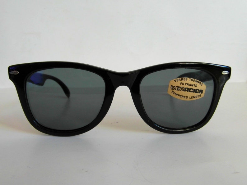 May include: Black plastic sunglasses with dark gray lenses. The sunglasses have a label that reads "VERRES TREMPES FILTRANTS UGRACIER TEMPERED LENSES".
