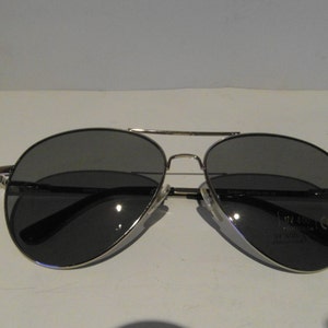 K&ouml;nnte beinhalten: Silberne Aviator-Sonnenbrille mit dunkelgrauen Gl&auml;sern. Die Sonnenbrille hat einen silbernen Metallrahmen und einen schwarzen Nasensteg.