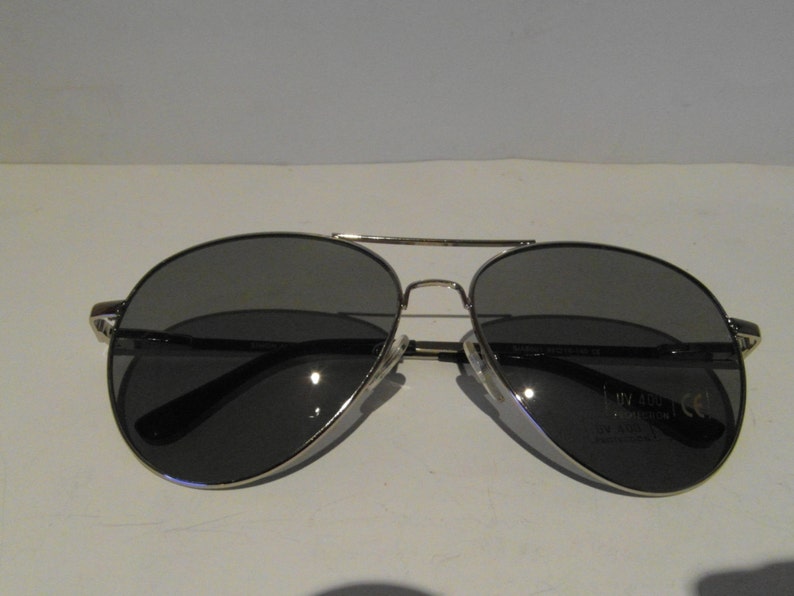 K&ouml;nnte beinhalten: Silberne Aviator-Sonnenbrille mit dunkelgrauen Gl&auml;sern. Die Sonnenbrille hat einen silbernen Metallrahmen und einen schwarzen Nasensteg.