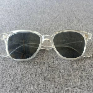 Peut inclure: Lunettes de soleil &agrave; monture transparente avec verres fonc&eacute;s. Les lunettes ont une forme classique et arrondie et sont fabriqu&eacute;es dans un mat&eacute;riau transparent. Les verres semblent &ecirc;tre gris fonc&eacute; ou noirs. La monture a une forme subtile et rectangulaire.