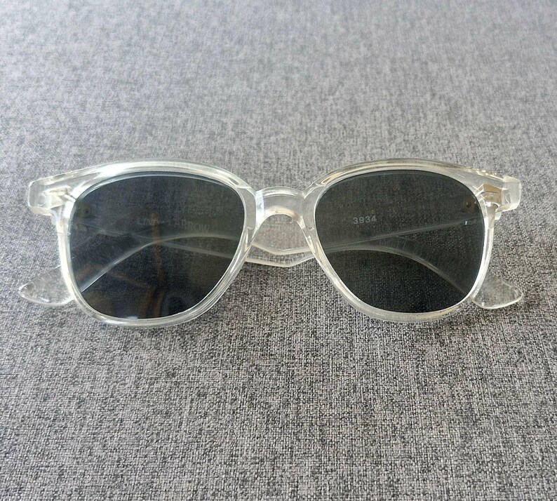 Peut inclure: Lunettes de soleil &agrave; monture transparente avec verres fonc&eacute;s. Les lunettes ont une forme classique et arrondie et sont fabriqu&eacute;es dans un mat&eacute;riau transparent. Les verres semblent &ecirc;tre gris fonc&eacute; ou noirs. La monture a une forme subtile et rectangulaire.