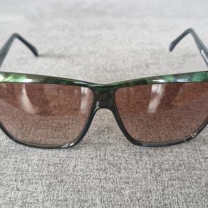 Puede incluir: Gafas de sol con montura verde oscuro y negra y lentes marrones. Las gafas de sol tienen una forma única y están sobre un fondo gris.