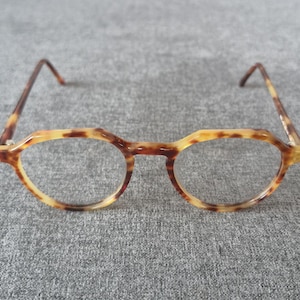 以下が含まれることがあります： べっ甲柄の眼鏡。フレームは丸みを帯びた形状で、幾何学的な上部があります。テンプルとブリッジは濃い茶色です。レンズは透明です。