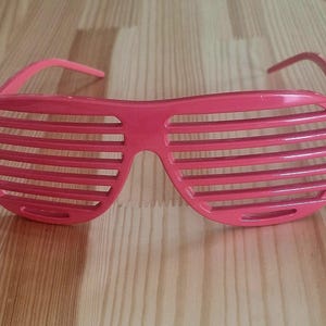 May include: Bright pink shutter-style sunglasses with horizontal slats across the lenses. The sunglasses are made of plastic and have a retro, 80s aesthetic. The frames and arms are a solid, vibrant pink color.