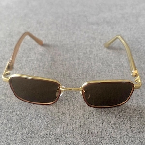 May include: Rectangular sunglasses featuring a thin gold-tone metal frame and dark brown tinted lenses. The temples are a translucent light brown material, and the bridge connects the two lenses with a simple, straight gold-tone metal bar.