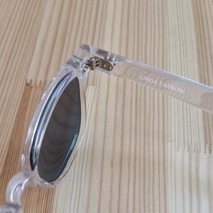 Peut inclure: Lunettes de soleil transparentes avec verres fonc&eacute;s. La monture est transparente et le texte "LINDA FARROW" est imprim&eacute; sur la branche. La charni&egrave;re est visible, et les lunettes reposent sur une surface en bois clair.