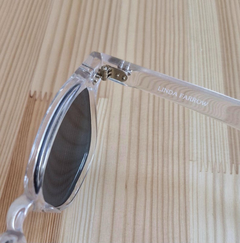 Peut inclure: Lunettes de soleil transparentes avec verres fonc&eacute;s. La monture est transparente et le texte "LINDA FARROW" est imprim&eacute; sur la branche. La charni&egrave;re est visible, et les lunettes reposent sur une surface en bois clair.