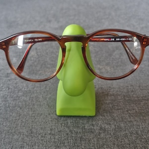 May include: Brown tortoise shell round glasses with TRUDEL-BLOG and 50-20-145 printed on the frame, sitting on a green plastic nose shaped glasses holder.