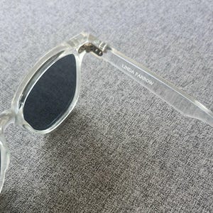 Peut inclure: Lunettes de soleil transparentes avec verres fonc&eacute;s. La monture est transparente et le texte "LINDA FARROW" est imprim&eacute; sur la branche. Les lunettes sont inclin&eacute;es, montrant la charni&egrave;re et la forme des verres.