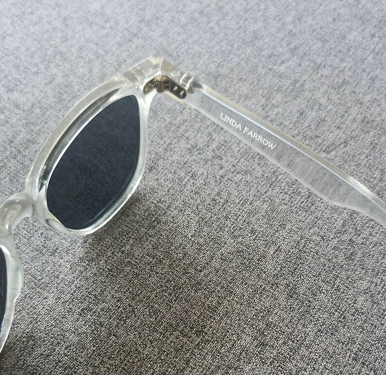 Peut inclure: Lunettes de soleil transparentes avec verres fonc&eacute;s. La monture est transparente et le texte "LINDA FARROW" est imprim&eacute; sur la branche. Les lunettes sont inclin&eacute;es, montrant la charni&egrave;re et la forme des verres.