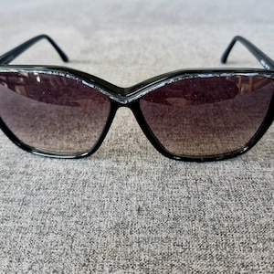 Puede incluir: Gafas de sol negras con forma de ojo de gato. Las lentes tienen un degradado de oscuro a claro. Las monturas negras tienen un detalle plateado en el borde superior. Las patillas son finas y negras.