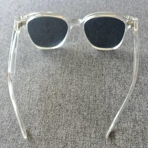 Peut inclure: Lunettes de soleil en plastique transparent avec des verres fonc&eacute;s. Les lunettes ont une forme rectangulaire classique avec une monture et des branches transparentes. Les verres sont d'une couleur fonc&eacute;e unie, offrant de l'ombre et une protection solaire.