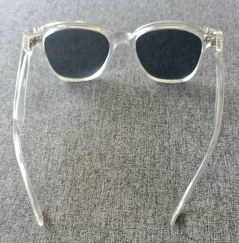 Peut inclure: Lunettes de soleil en plastique transparent avec des verres fonc&eacute;s. Les lunettes ont une forme rectangulaire classique avec une monture et des branches transparentes. Les verres sont d'une couleur fonc&eacute;e unie, offrant de l'ombre et une protection solaire.